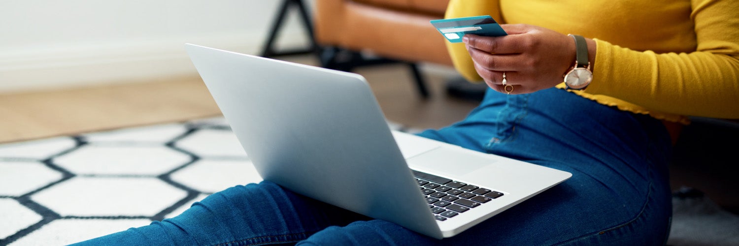 Woman sitting by the sofa shopping online on laptop with bank card in hand. 