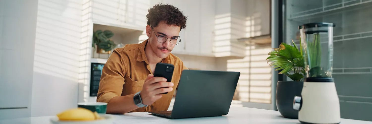 Person sitting at a kitchen table on the phone, reviewing paperwork next to an open laptop.